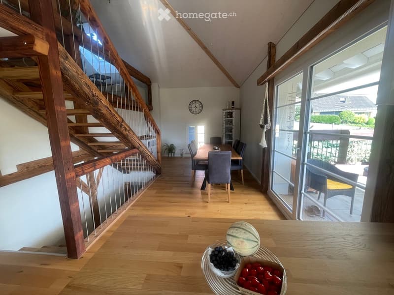 Bright, open-plan living and dining area with wooden flooring, a staircase leading upstairs, and large windows offering a view of the garden. The dining area features a wooden table with chairs, and a fruit basket is placed on the coffee table.