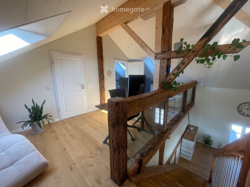 Bright, wooden-floored room with a staircase leading to an upper level, featuring a sofa, a small table with a plant, and a workspace with a screen and chair.