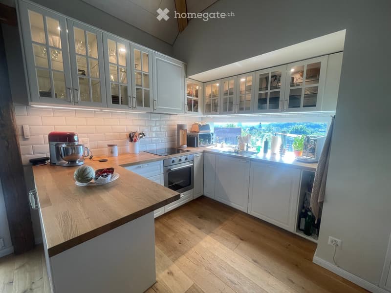 Modern kitchen with white cabinets, wooden countertop, and stainless steel appliances. Features an island with a mixer, bowl, and fruit, and a window overlooking a garden.