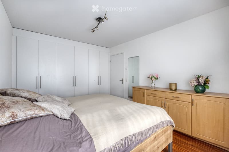 Modern bedroom with a large bed, wooden wardrobe, dresser with vases, and a mirror on the wall.