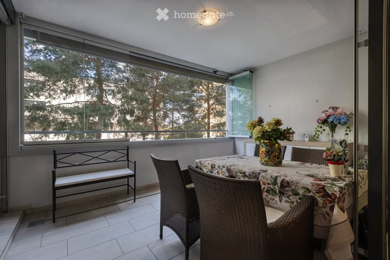 Balcony with a dining table, chairs, a bench, and vases with flowers, overlooking a tree-lined area.