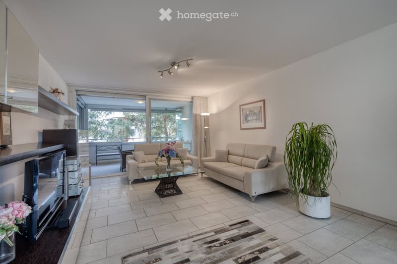 Modern living room with two beige sofas, a glass coffee table, a potted plant, and a TV on a stand. Large windows offer a view of the balcony and outdoor area.