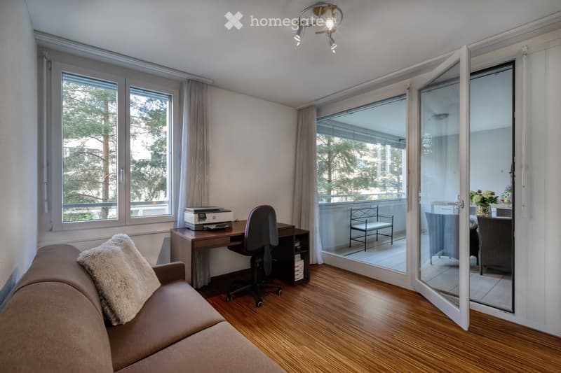 Modern furnished room with a sofa, office chair, desk, printer, and a view of a balcony area with outdoor furniture.