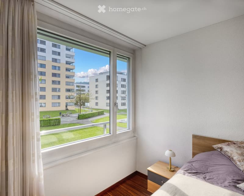 Bright bedroom with wooden flooring, large window offering a view of a green park and modern buildings, featuring a bed with pillows, a nightstand, and a lamp.