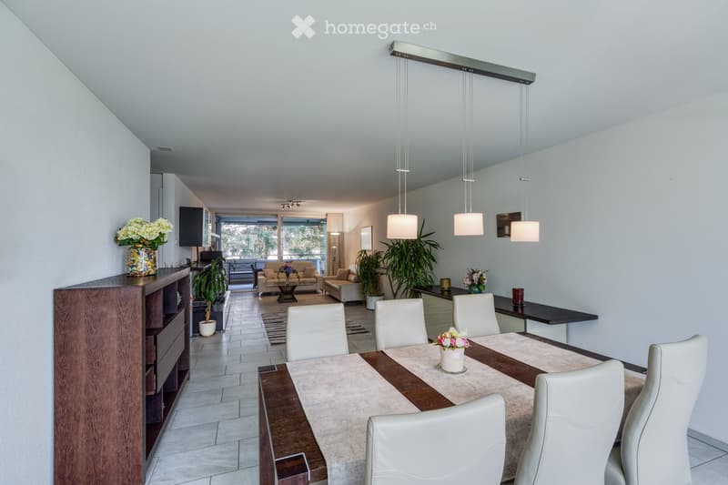 Modern dining area with a wooden dining table, white chairs, and a beige table runner. A living area with a beige sofa and coffee table is visible in the background, along with potted plants and decorative vases.