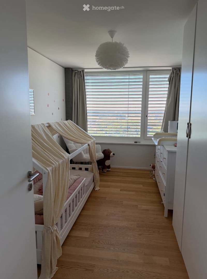 Bright, modern children's bedroom with a wooden floor, large window with blinds, a white bed with a beige canopy, a dresser, and a plush toy on the floor.