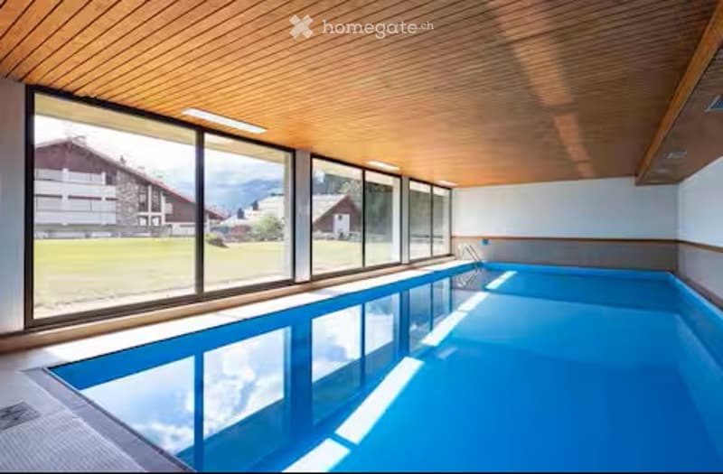 Indoor swimming pool area with large windows offering a scenic mountain view, wooden ceiling, and bright lighting.