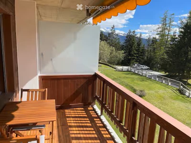 Balcony with wooden floor, railing, table, and chairs, overlooking a green lawn and mountain view.