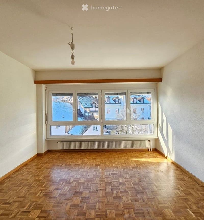 Empty room with wooden flooring, large windows showing a cityscape, and a radiator below the windows.