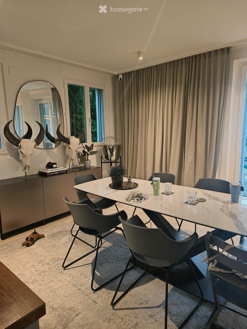 Modern dining area with a white marble table, black chairs, a large mirror, a cabinet with a skull decor, and potted plants.