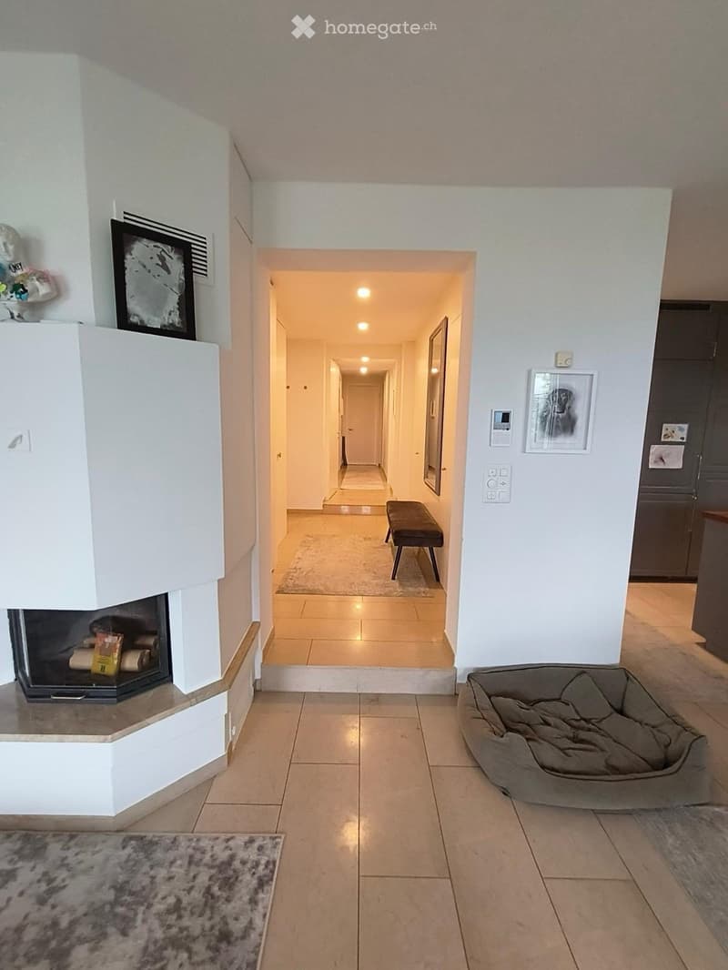 Bright hallway with tiled flooring, white walls, and a fireplace on the left. A small bench is placed in the adjacent room, and a pet bed is visible on the right side.