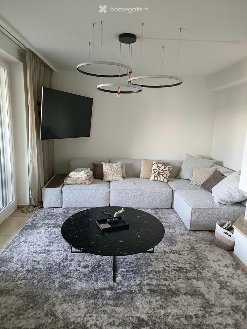 Modern living room with a grey sectional sofa, decorative pillows, a black coffee table, a large television mounted on the wall, and a grey shag rug.