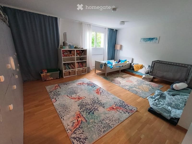 Bright, child-friendly bedroom with wooden flooring, large area rug, bed with pillows and blankets, shelf with toys and books, and a small seating area with cushions.