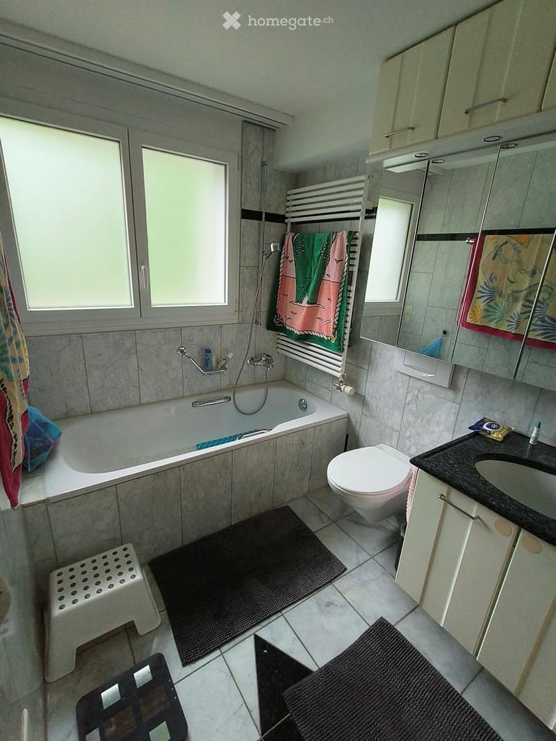 Bathroom with a bathtub, toilet, sink, mirror, and cabinets. Includes a towel rack with towels, a small stool, and bathmats on the floor.