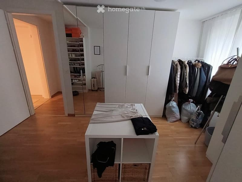 A well-lit, organized walk-in closet with wooden flooring, large white wardrobe, a small table with folded clothes, and a clothing rack with hanging garments.