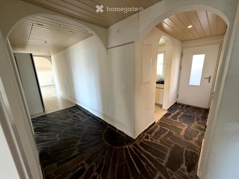 A narrow hallway with white walls, stone flooring, and arched doorways leading to other rooms. The ceiling is light-colored with recessed lighting.
