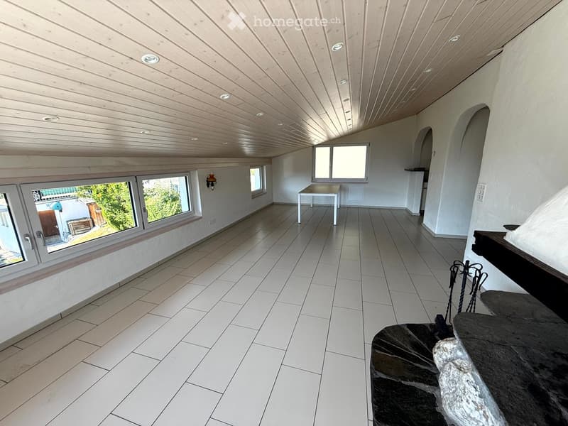 Bright, open room with white tiled flooring, large windows, and a slanted wooden ceiling. Minimal furnishings include a small table and a stone bench.