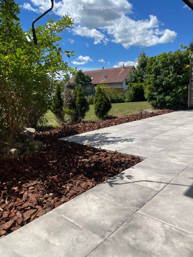 Garden area with a concrete pathway bordered by mulch and small rocks, with a house and trees visible in the background.