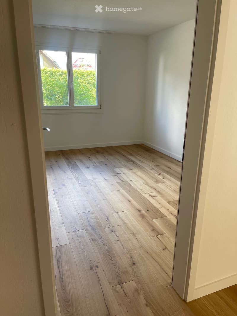 Empty room with light wooden flooring and white walls, featuring a large window with a view of greenery outside.