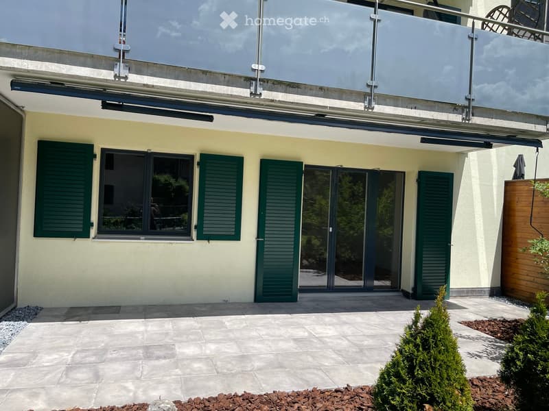 Modern building exterior with large windows, green shutters, and a glass balcony overlooking the street.
