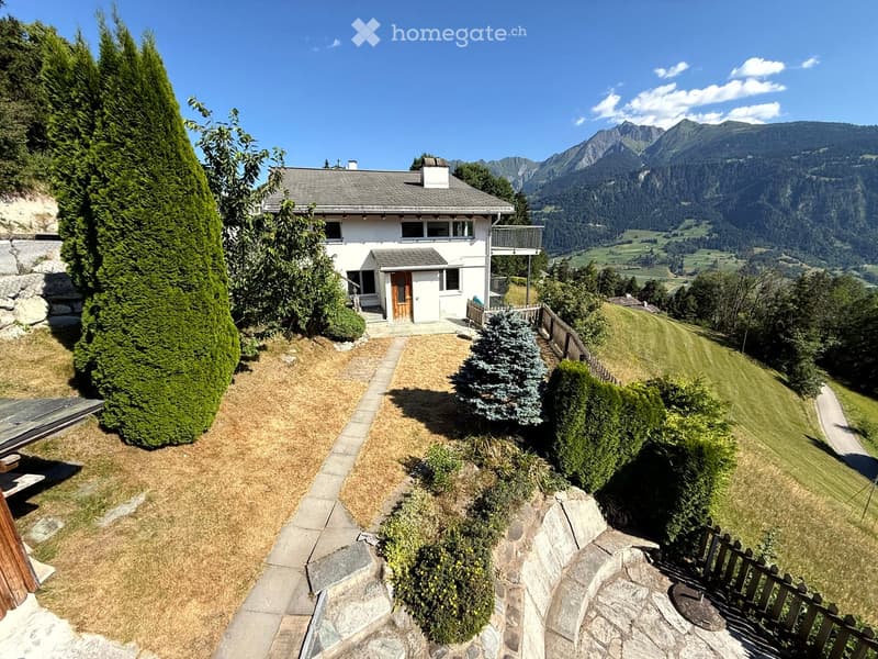 A two-story white house with a gray roof, situated on a hillside with a scenic mountain view in the background. The house features a wooden front door, multiple windows, and a small balcony on the upper level. The surrounding area includes a well-maintain