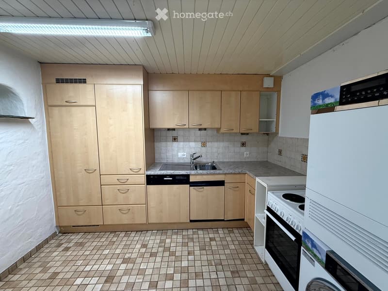 A well-lit kitchen with light wooden cabinets, tiled walls, and a tiled floor. The kitchen features a sink, dishwasher, oven, refrigerator, and microwave.