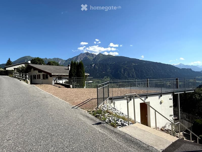 A two-story house with a sloped roof and white exterior, situated on a raised ground floor with a gravel driveway and a metal staircase leading to a balcony overlooking a scenic mountain view.