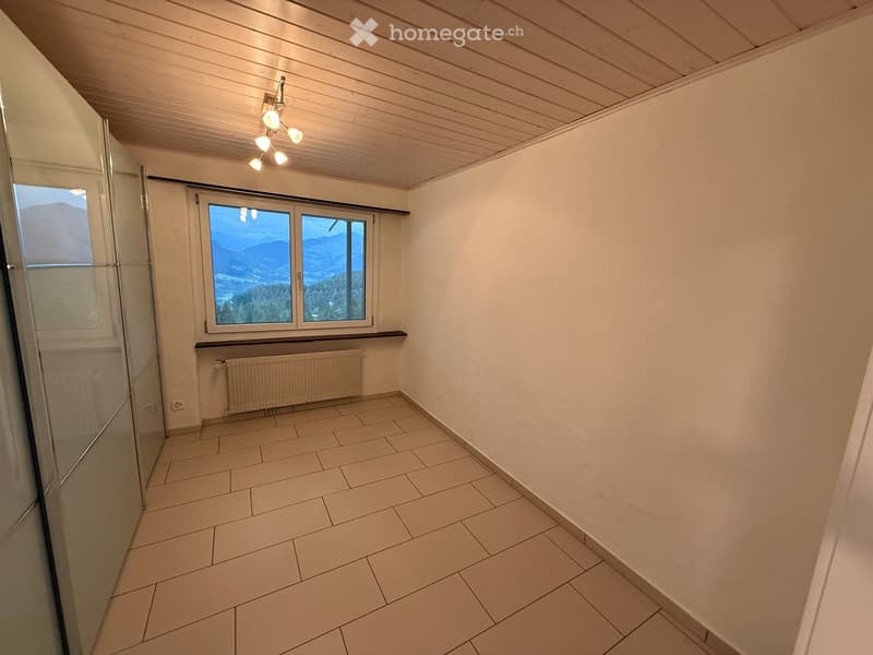Empty room with tiled floor, white walls, wooden ceiling, and large window offering a mountain view. Includes a radiator and a glass shower enclosure visible on the left side.