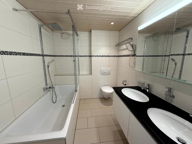 Modern bathroom with white tiles, a bathtub, toilet, double sink, and a large mirror. Features a glass-enclosed shower area with a handheld showerhead.