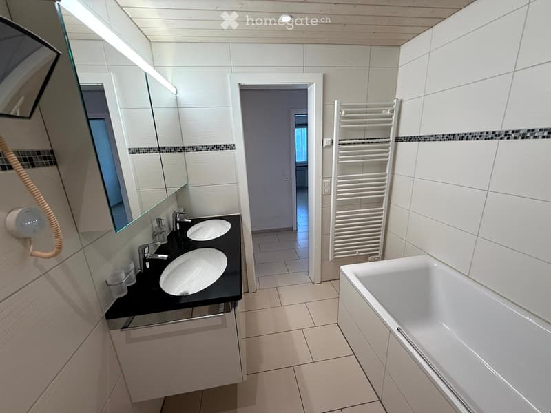 Modern bathroom with white tiles, double sink, bathtub, and large mirror. Features a towel rack and a built-in storage cabinet under the sink.