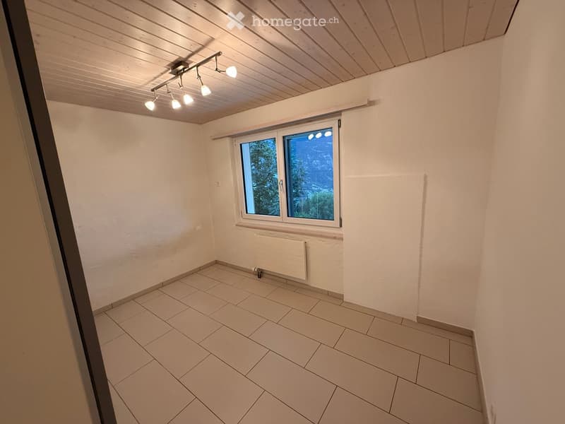 Empty room with tiled floor, white walls, wooden ceiling, and a window showing a view of trees.
