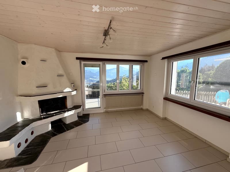 Empty room with white tiled floor, white paneled ceiling, large windows with mountain view, fireplace with built-in seating, radiator, and modern lighting fixtures.