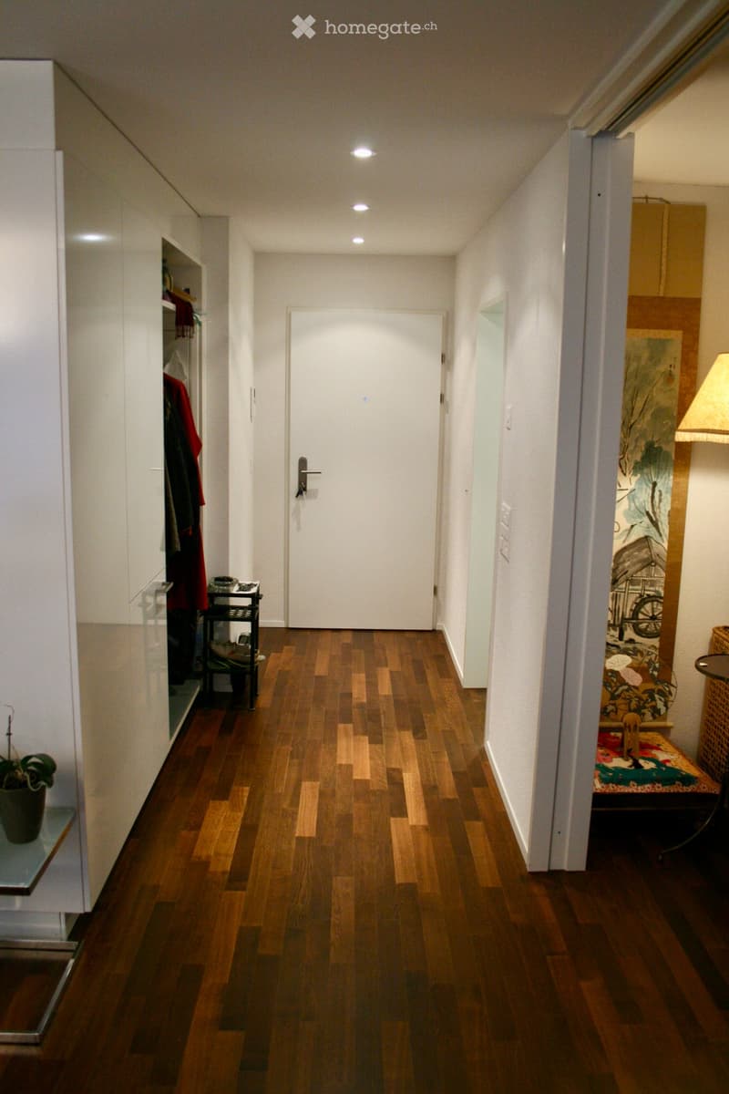 A narrow hallway with wooden flooring, white walls, and recessed ceiling lighting. Features a white door at the end, a small shelf with a plant, and a small table with items on the right side.