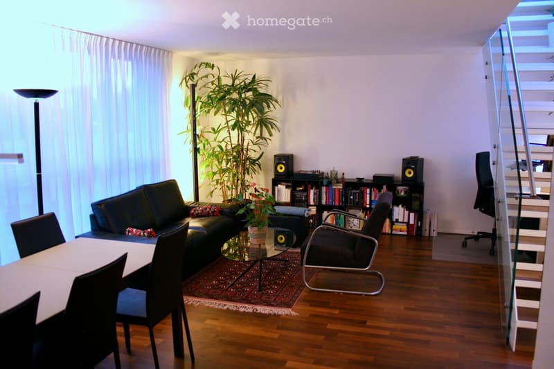 A modern living room with a black leather sofa, glass coffee table, dining table with chairs, bookshelf, and a large potted plant. The room features wooden flooring and a glass staircase leading to an upper level.
