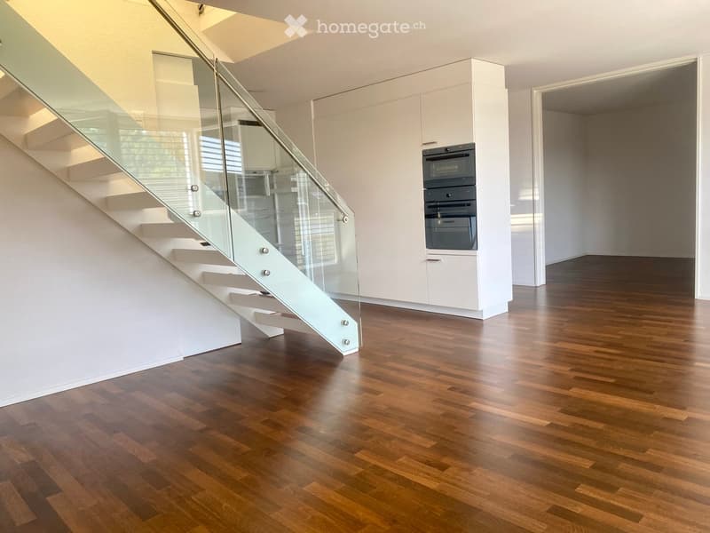 Modern, minimalist interior with polished wooden flooring, white walls, and a sleek staircase with glass railing leading upwards. Built-in cabinets and appliances are visible in the background.