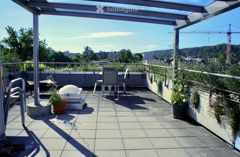 A modern rooftop terrace with a glass roof, tiled floor, and outdoor furniture including chairs, a table, and potted plants. The area features a railing and offers a view of the city and surrounding greenery.