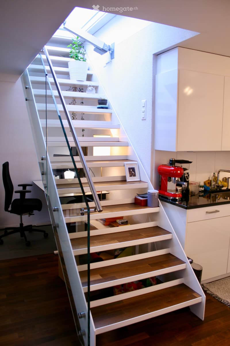 Modern, compact room with a staircase leading up, featuring a desk with a chair, a coffee maker on a counter, and a small plant on the stairs.