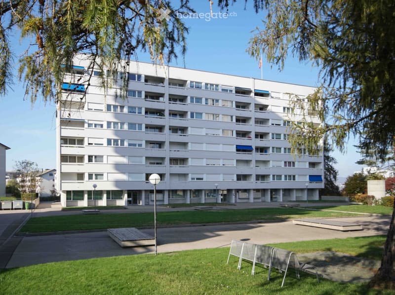 Multi-story residential building with numerous windows and balconies, surrounded by a landscaped area with grass, trees, and outdoor seating.