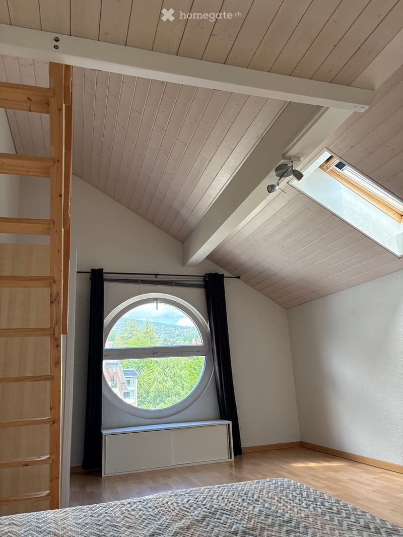 Bright bedroom with wooden ceiling and floor, large round window offering a scenic view, bed with patterned bedding, built-in cabinet, and black curtains.