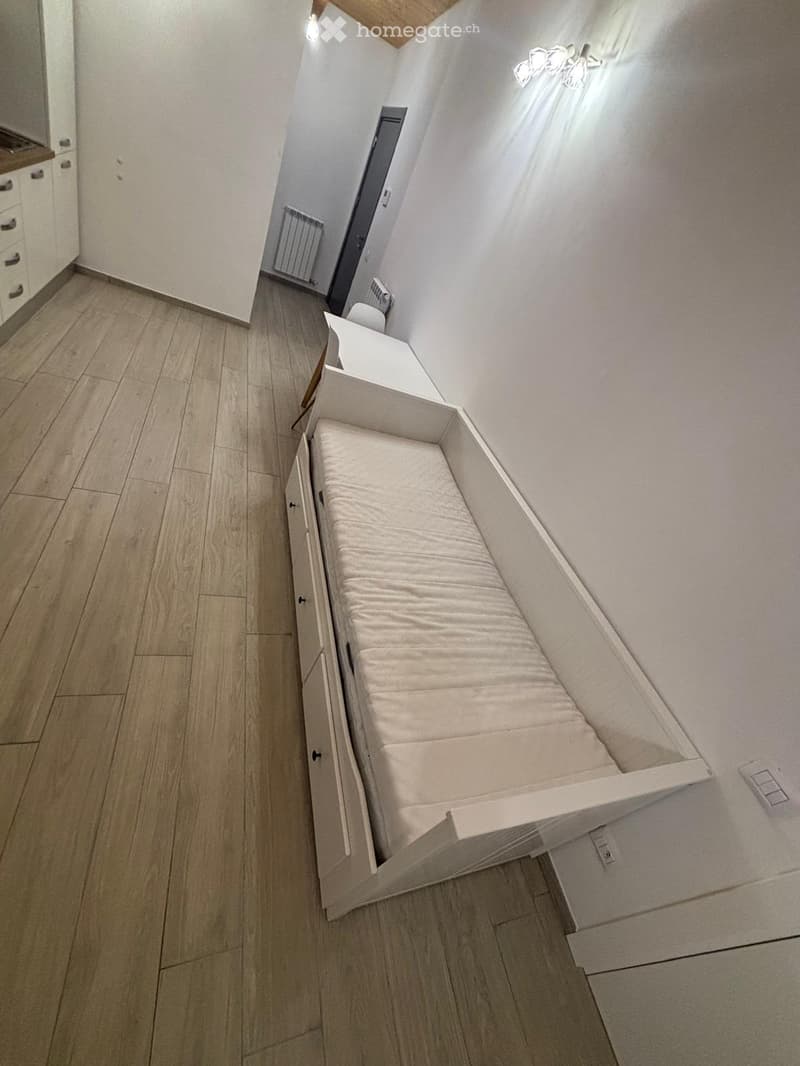 A modern, compact bedroom with a built-in sofa bed and a small cabinet, featuring light wooden flooring and white walls.