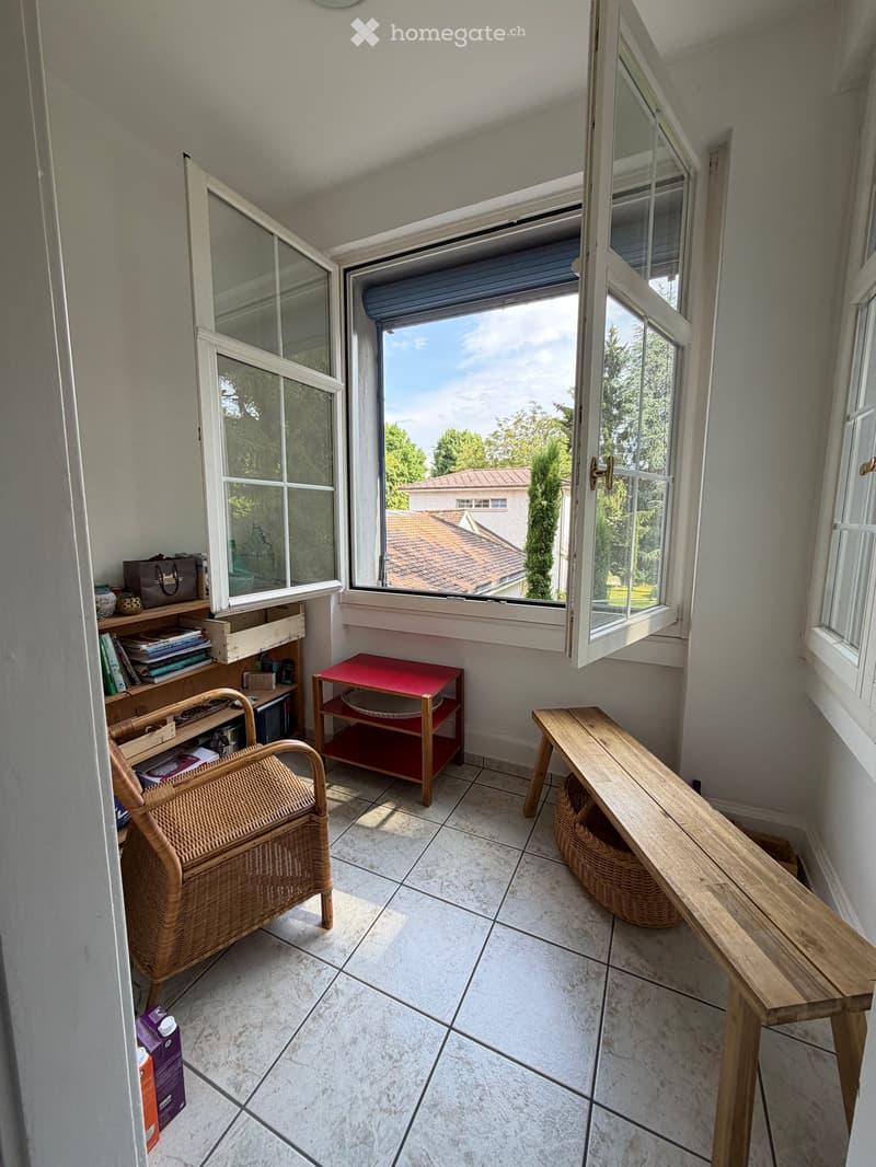 Balcony with tiled floor, large windows, wooden bench, wicker chair, small red table, shelf with books and items.
