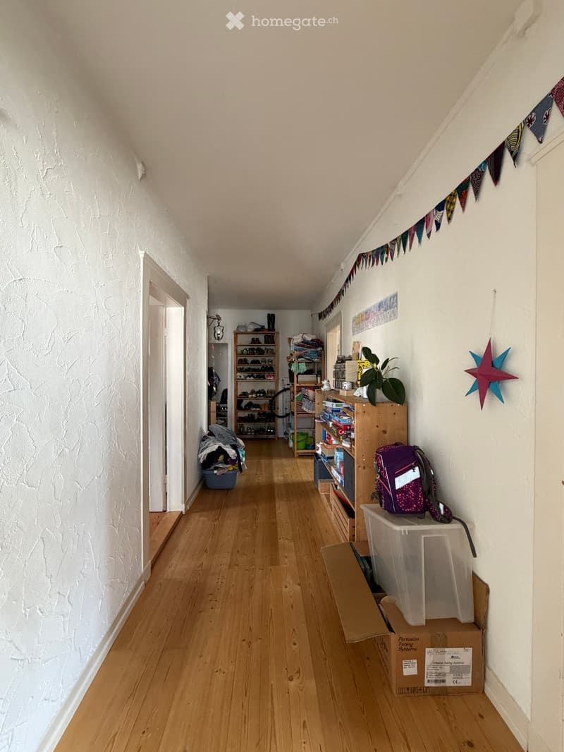 A narrow hallway with wooden flooring, white walls, and various storage items including shelves, boxes, and a backpack. The hallway has a slanted ceiling and a colorful bunting hanging on one wall.