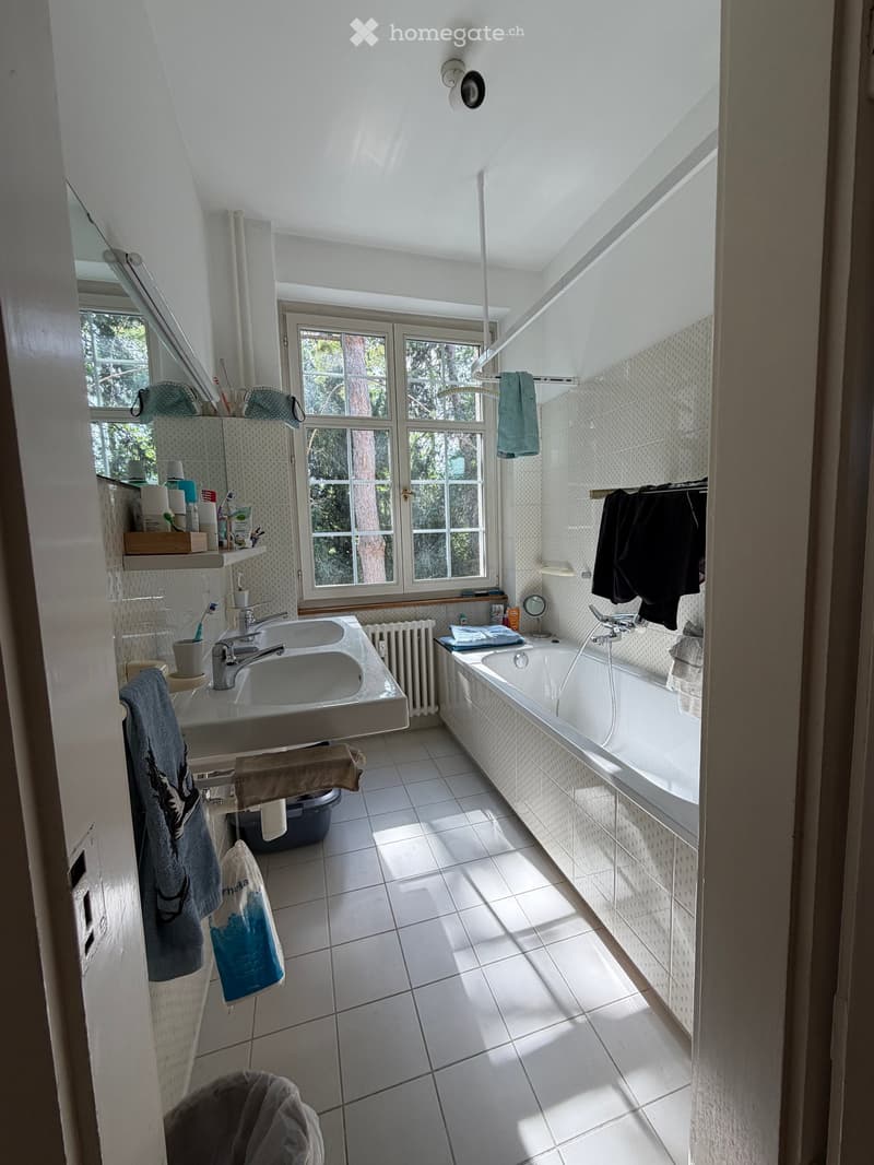 Bright bathroom with white tiled floor and walls, double sink, bathtub, large window, and various personal items on shelves and surfaces.