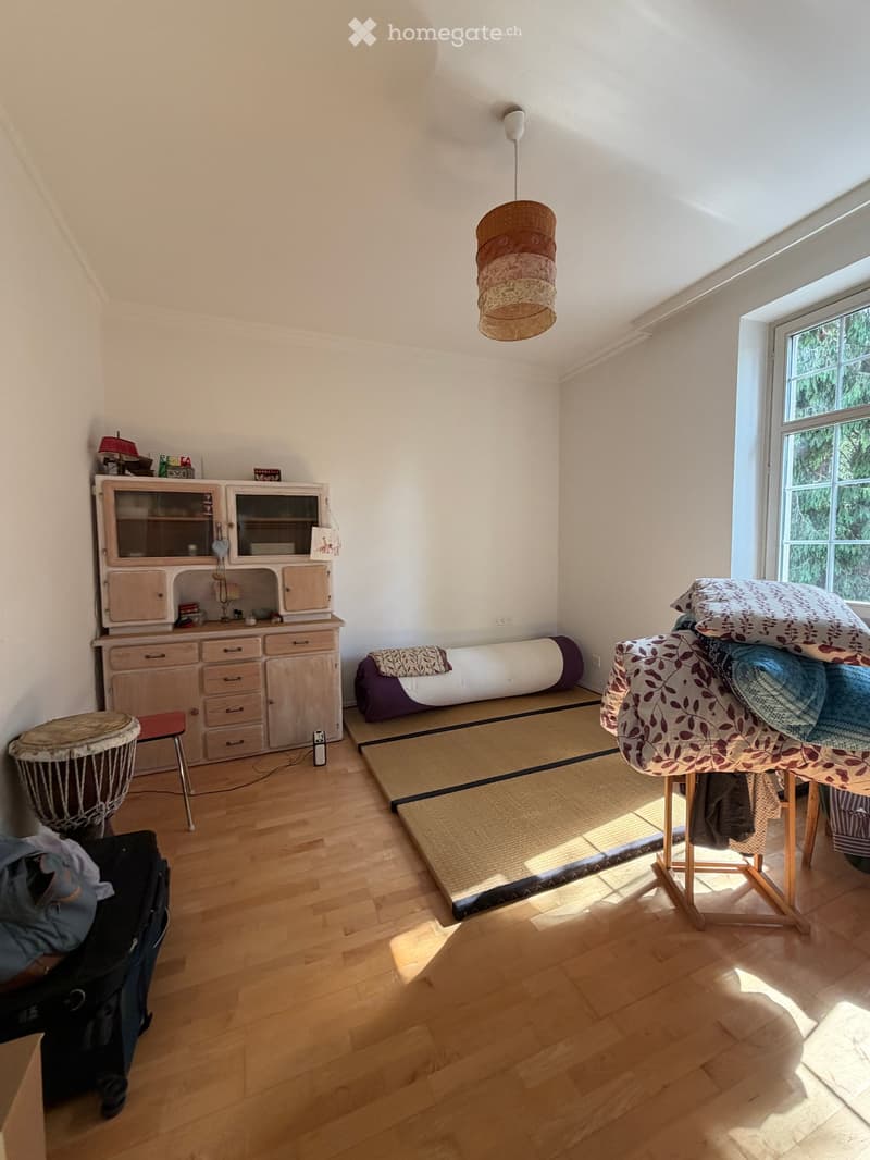 Bright room with wooden flooring, large window, cabinet, stool, drum, table with pillows and blankets, yoga mat, and yoga bolster.