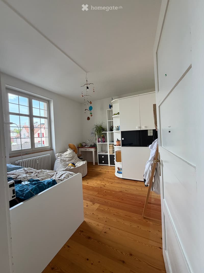 Bright bedroom with wooden flooring, large window, bed with blue bedding, chair, desk, bookshelf, and cabinet.
