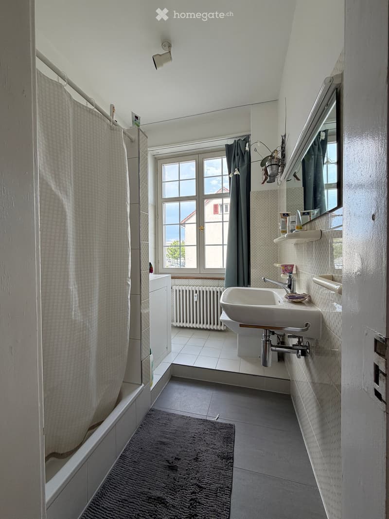 Bathroom with a sink, mirror, bathtub, and window with curtains. Includes a radiator and a small rug on the floor.