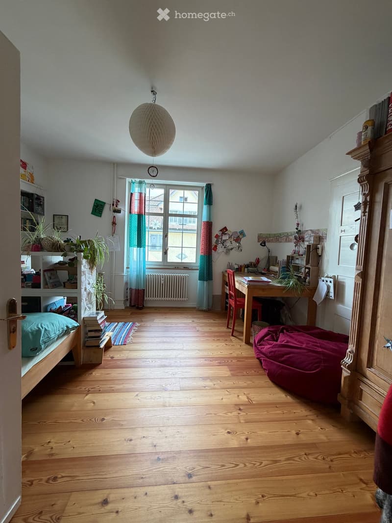 A well-lit bedroom with wooden flooring, a bed with a teal pillow, a desk with a chair, a beanie chair, shelves with books and plants, and a window with colorful curtains.