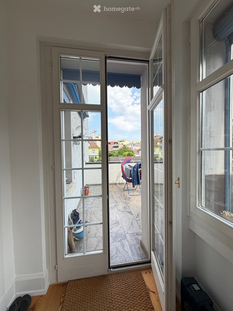 Hallway with wooden flooring, carpeted area, and an open sliding door leading to a tiled balcony with outdoor items like shoes, a broom, and a chair.