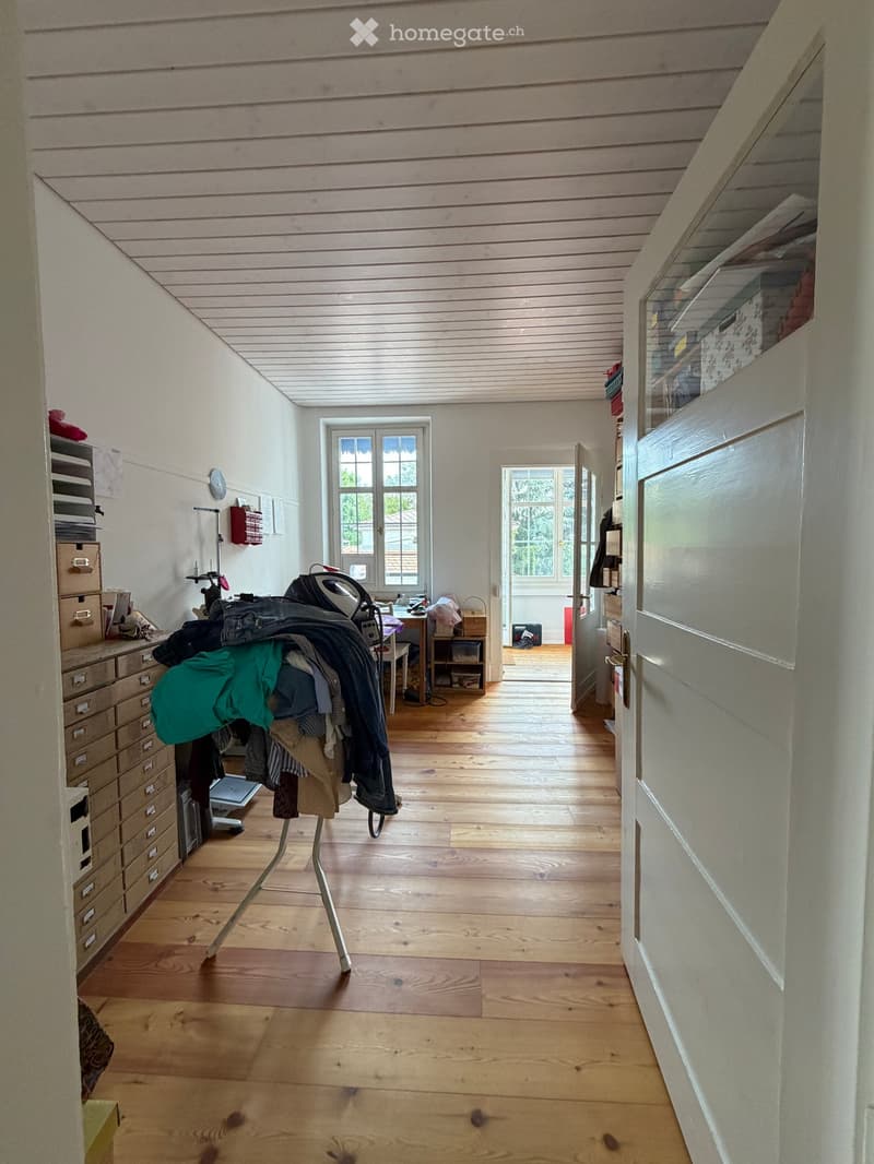 A well-lit room with wooden flooring, a slanted ceiling, and large windows. Contains a desk with a chair, a clothing rack with folded clothes, a cabinet, and a table with items on it.