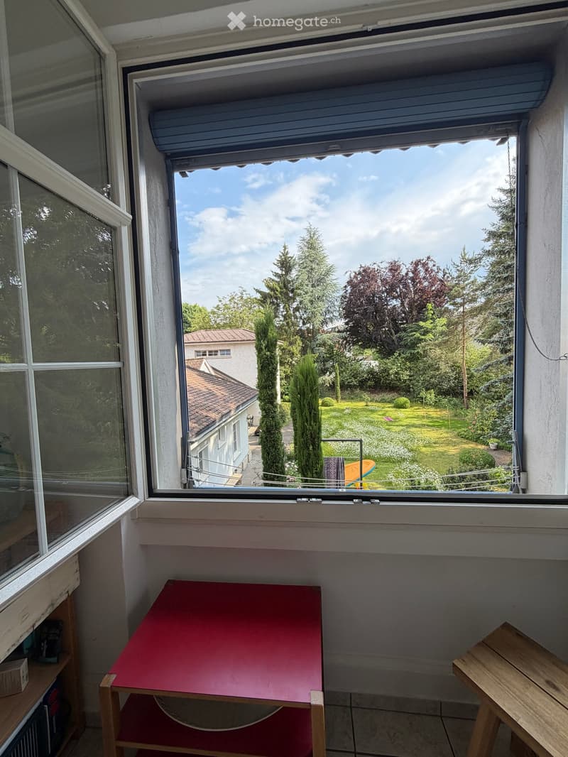 A small room with a red table, wooden stool, and shelf, with a large window offering a view of a garden and neighboring house.
